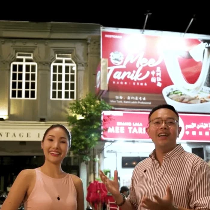 Two hosts presenting at night outside a lit Mee Tarik noodle shop