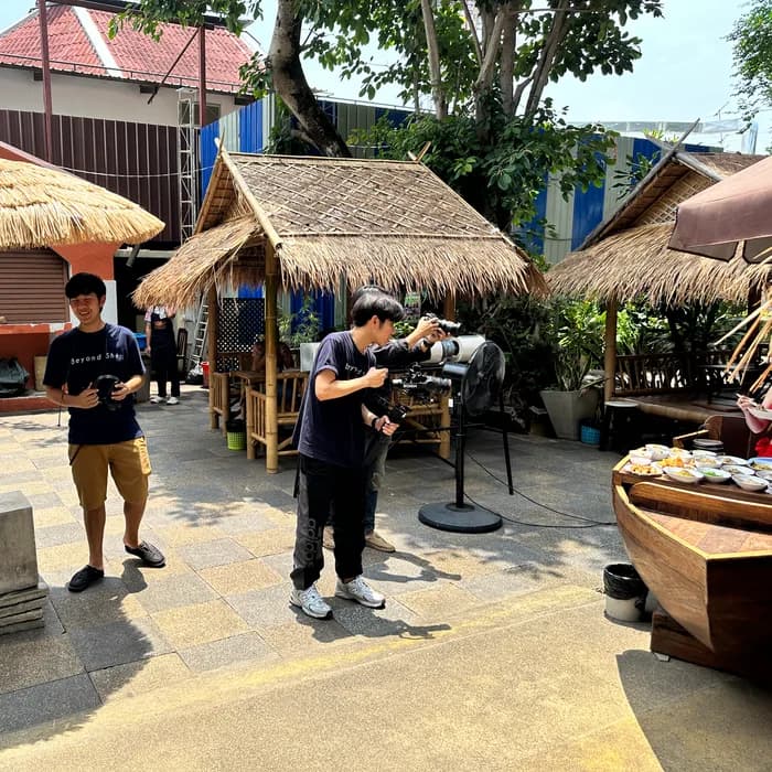 Crew setting up cameras around a boat table of Thai dishes in a thatched courtyard